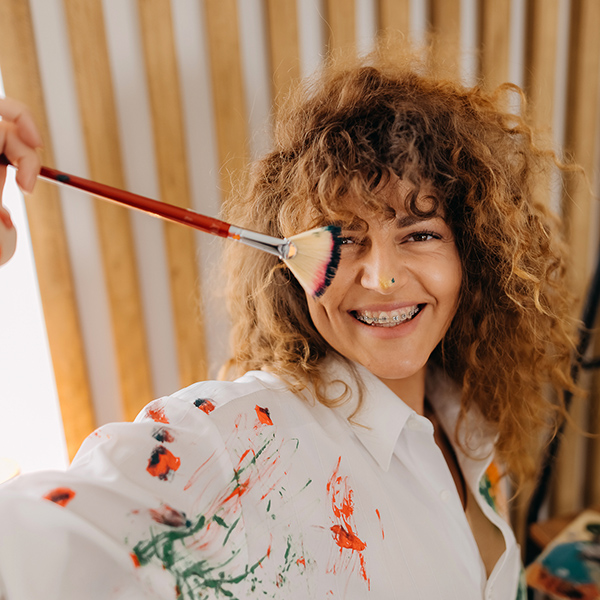 Female artist Lachende Dame mit Fächerpinsel in der Hand und Farbsprizern auf weißem Malhemd